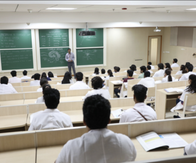 A medical classroom with students in white coats listening to a professor lecturing on the brain, with diagrams and notes on green chalkboards.